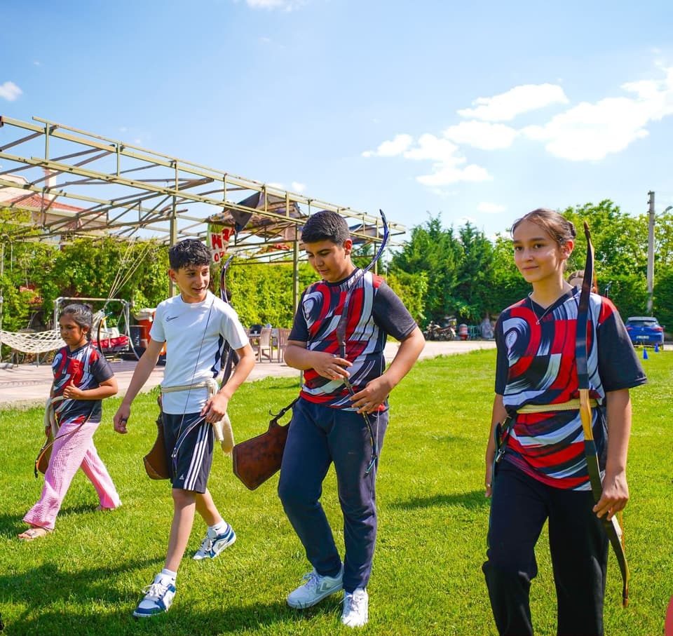 Geleneksel yaylarını taşıyan çocuklar çimenlikte atış hattına yürüyor