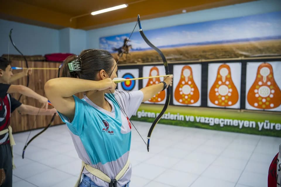 Kız sporcu salonda geleneksel yayla nişan alıyor; arkada puta hedefleri ve atlı okçu resmi