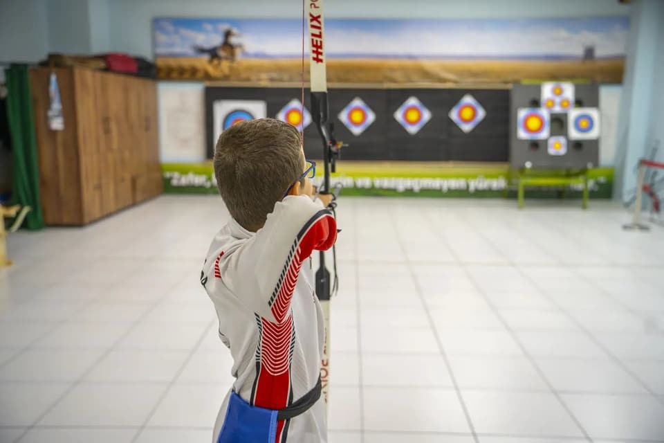 Salonda olimpik yayla atış yapan sporcu, arkadan; geniş atış alanı ve hedef duvarı