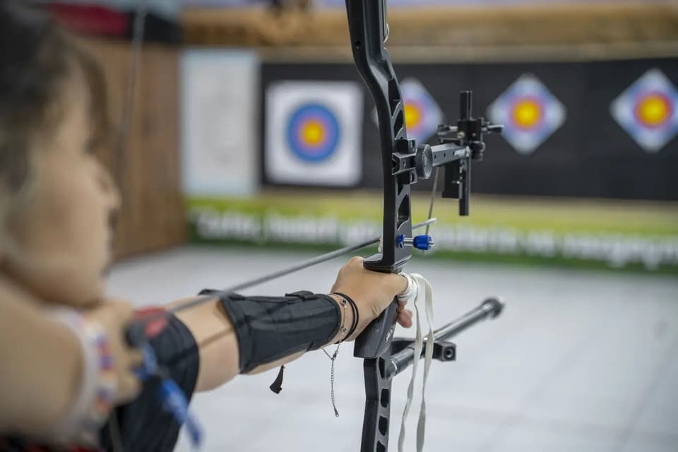Olimpik yayın nişangâhı ve stabilizatörü yakın planda; arkada hedefler bulanık